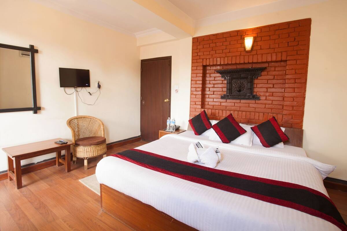 A hotel room with a large bed, a wooden bench, a wicker chair, a brick accent wall, and a flat-screen TV.
