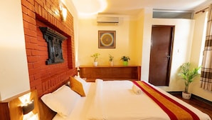 A hotel room with a brick accent wall, a bed with white linens, a wooden headboard, a bedside table with a lamp, and a potted plant on the windowsill. - Royal Atlantic Home  (Kathmandu)