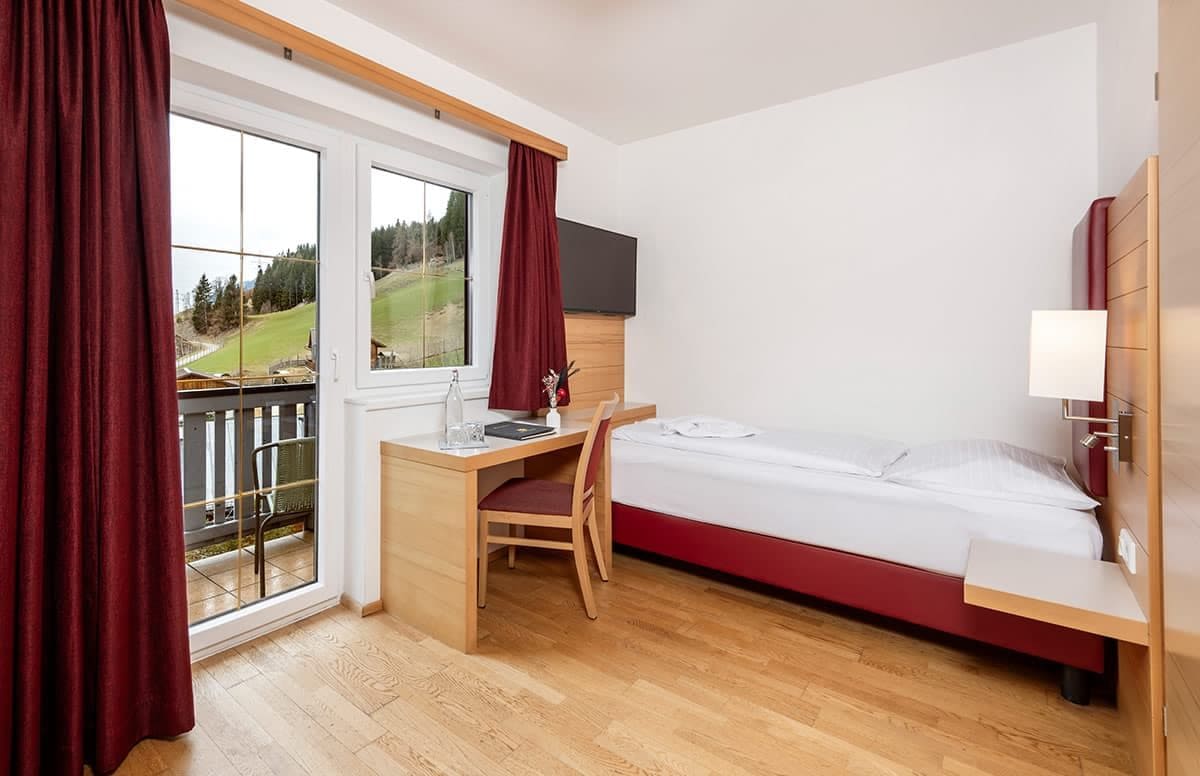 A hotel room with a bed, a desk, and a chair. There is a window with curtains, a TV mounted on the wall, and a balcony visible through the window.