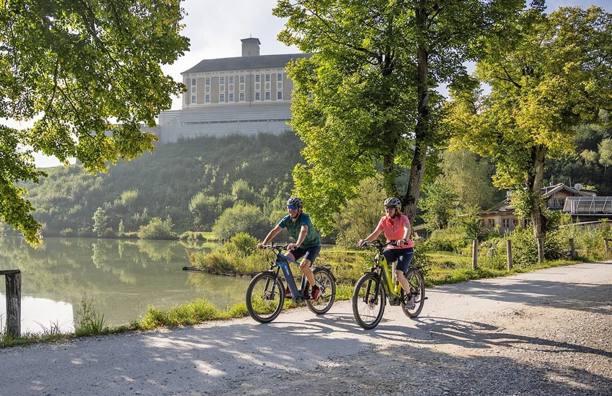 Dos ciclistas en un camino de grava con un cuerpo de agua y árboles al fondo.