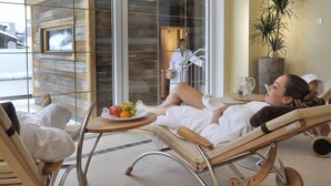 A spa suite with a glass door leading to a wooden cabin, a lounge chair, a small table with fruit, and a potted plant.