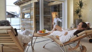 A room with a glass door leading to a balcony, a wooden-framed chair, a table with a fruit bowl, and a potted plant.