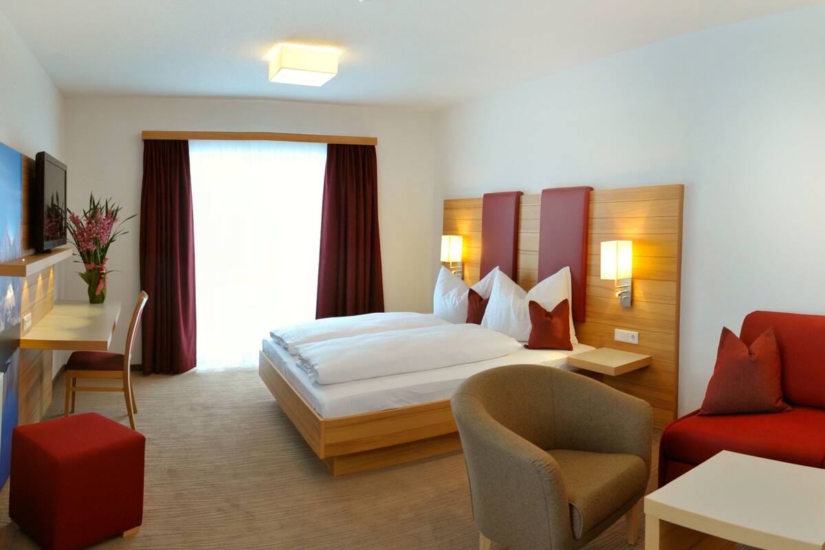 Une chambre d’hôtel avec un grand lit, deux tables de chevet avec des lampes, un bureau avec une chaise, un pouf rouge et un canapé rouge.