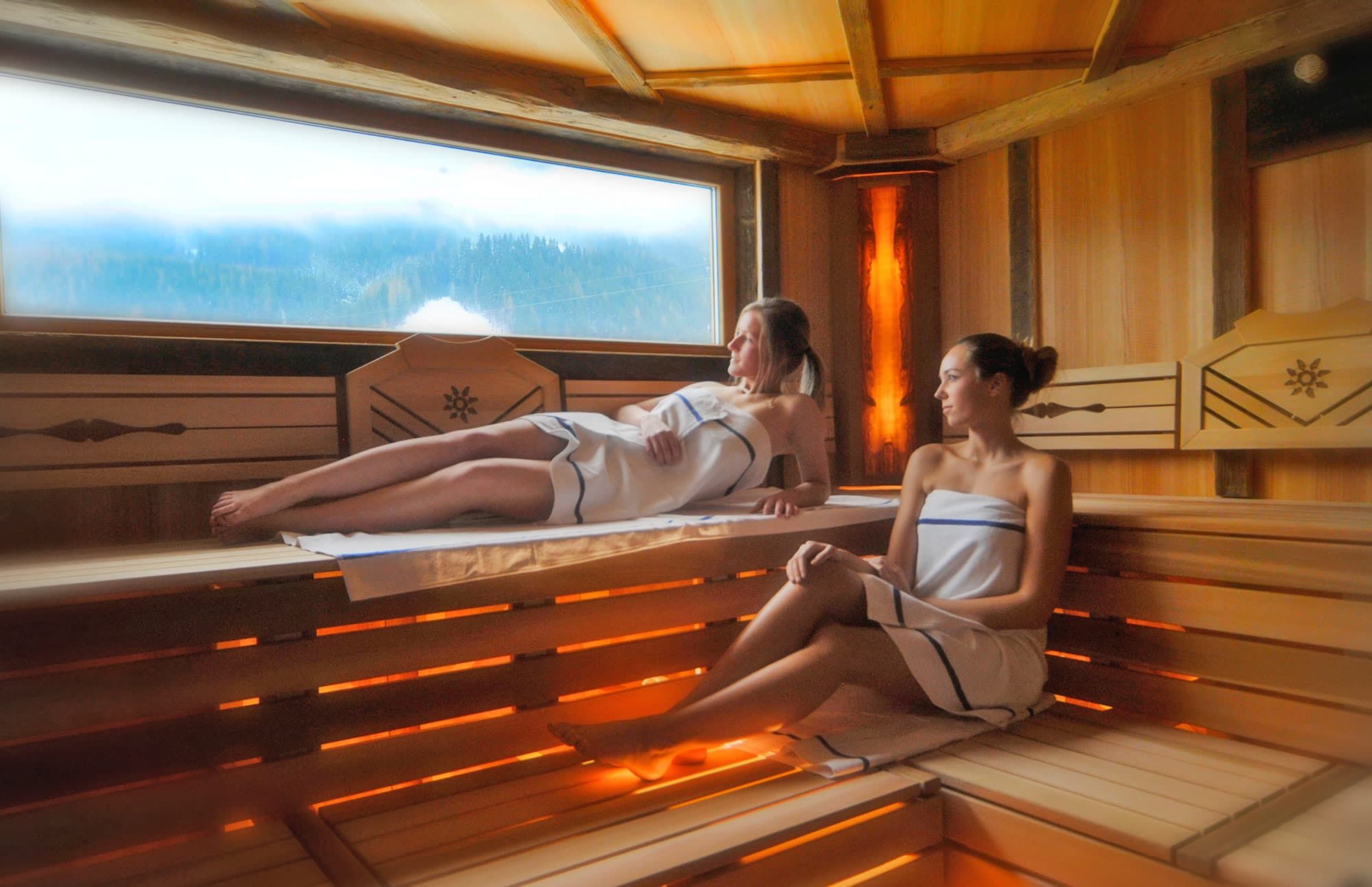 Two individuals in a sauna with wooden benches and a window overlooking a forested landscape.