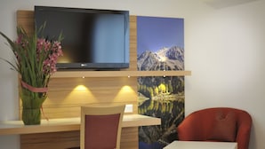 Ein Zimmer mit einem Holztisch, einem Stuhl, einem kleinen Tisch, einem an der Wand montierten Flachbildfernseher und einem Gemälde einer Berglandschaft.