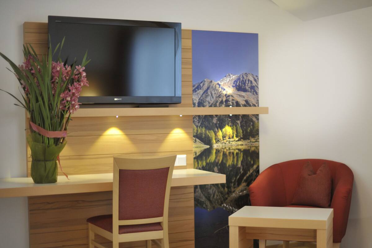 Une pièce comprenant un bureau en bois, une chaise, une petite table, un téléviseur à écran plat fixé au mur et un tableau représentant un paysage de montagne.