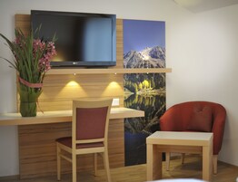 Una habitación con un escritorio de madera, una silla, una mesita pequeña, un televisor de pantalla plana montado en la pared y un cuadro de un paisaje montañoso.
