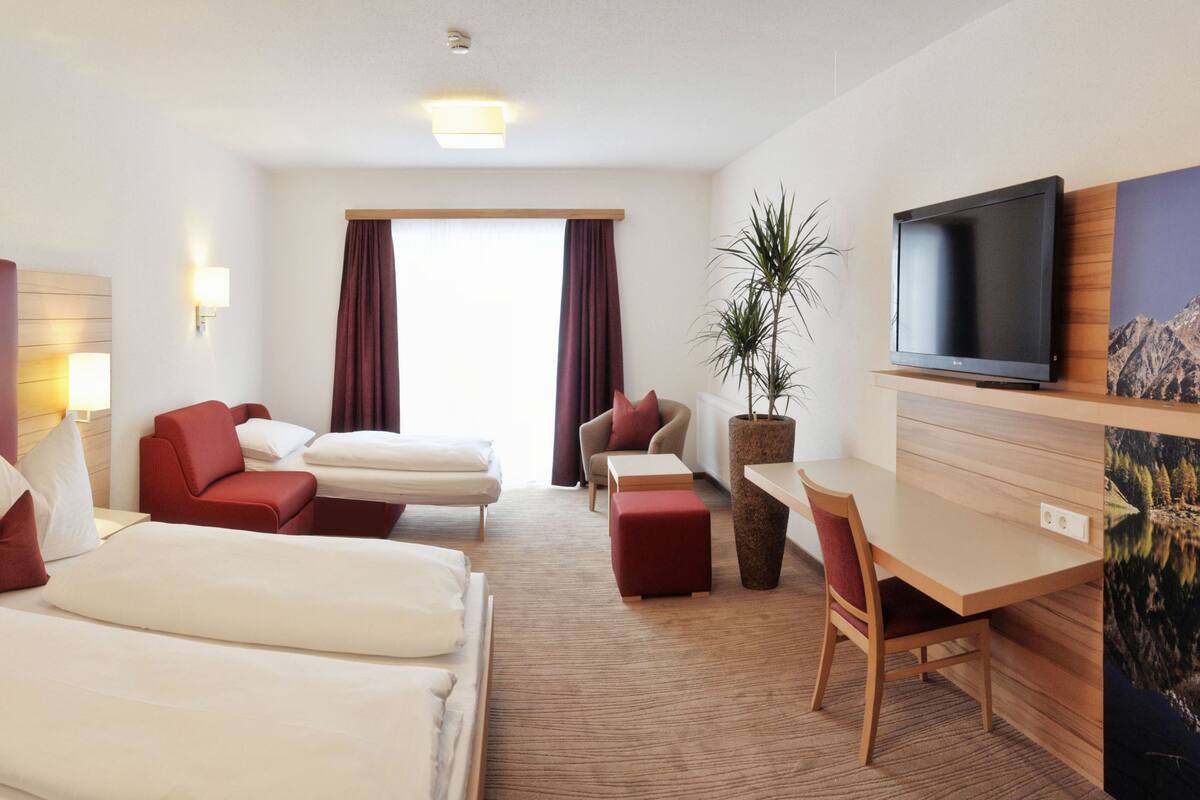 Une chambre d’hôtel avec un lit, deux fauteuils rouges, un canapé, un bureau, une télévision et une grande fenêtre avec des rideaux.