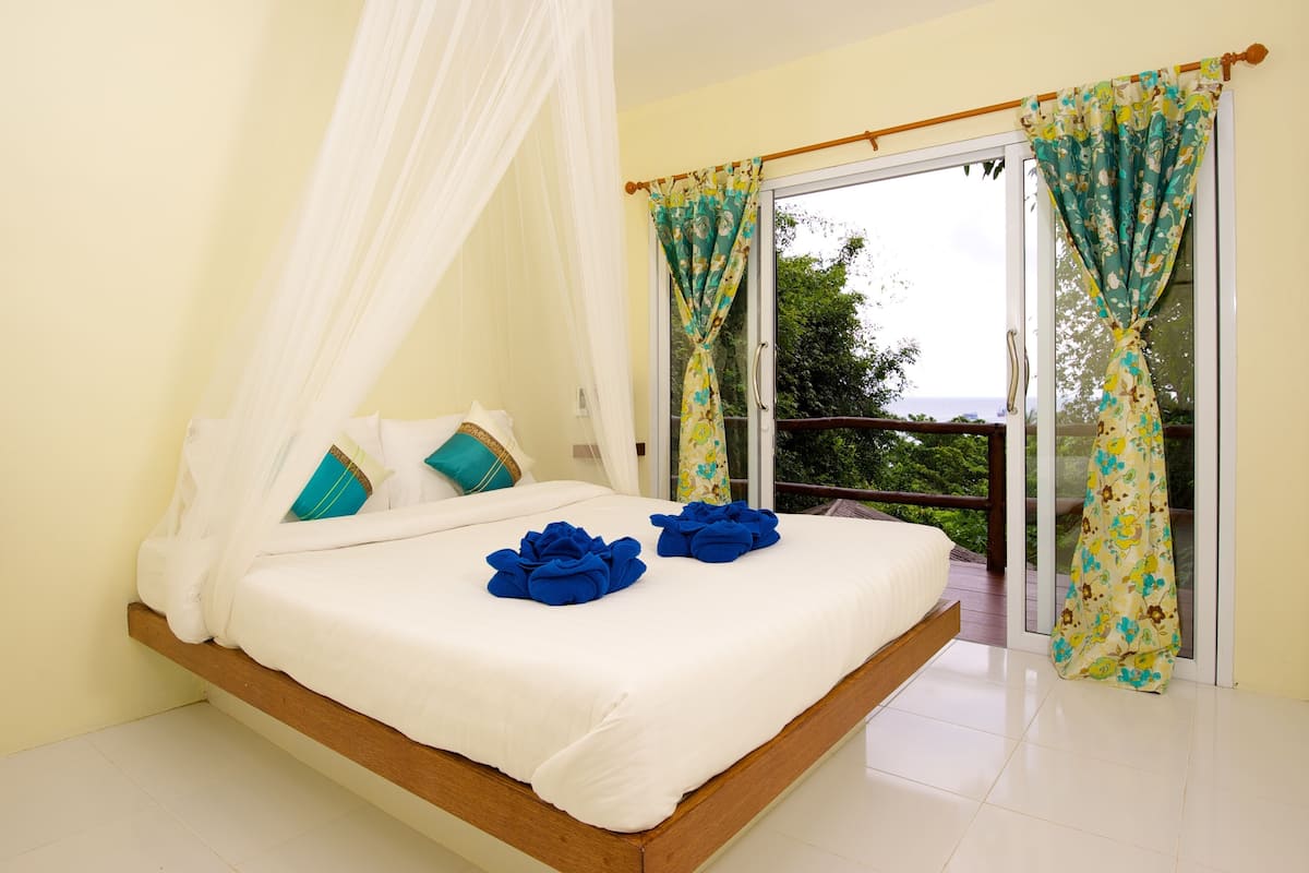 A bedroom with a bed, white curtains, and a balcony with a view.