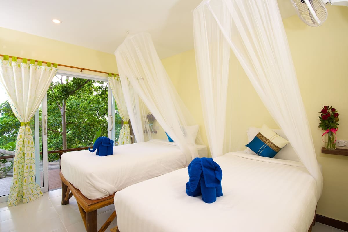 A room with two beds, each with a canopy. There is a window with curtains, a fan, and a vase with flowers on a nightstand.