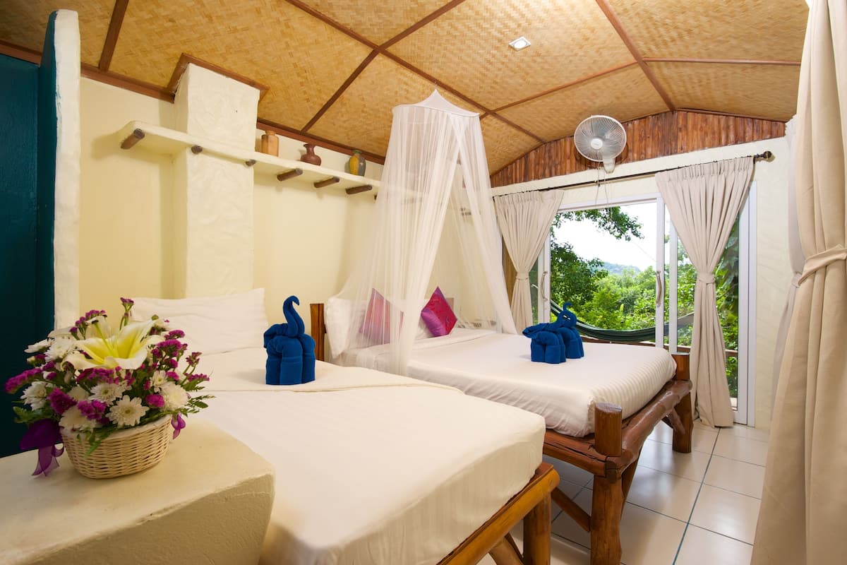 A room with two beds, a ceiling fan, a window with curtains, and a basket of flowers.