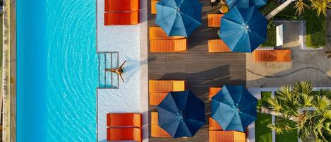 An aerial view of a pool area with multiple orange lounge chairs and blue umbrellas, surrounded by a wooden deck and palm trees.