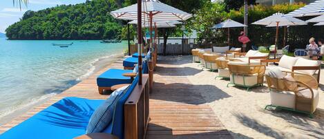 Una terraza de madera con asientos acolchados azules y sombrillas a rayas, con vistas a una playa de aguas turquesas cristalinas y una isla verde exuberante al fondo.