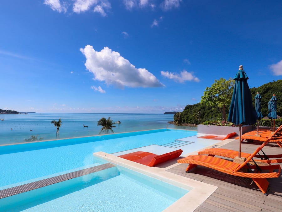 Piscina azul límpida com espreguiçadeiras e guarda-sóis, com vista para um mar calmo e vegetação exuberante.