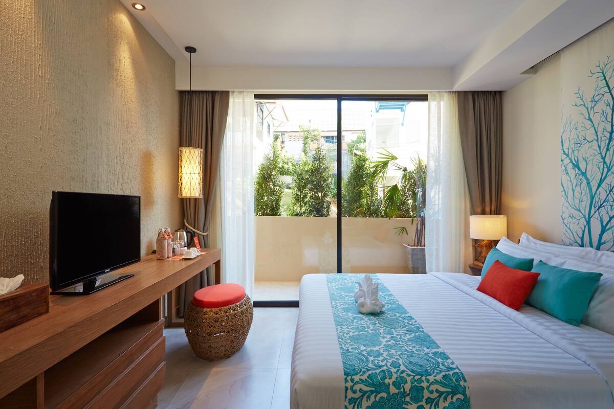 A hotel room with a flat-screen TV, a wooden side table, a bed with patterned bedding, a red pillow, and a view of greenery through the balcony doors.
