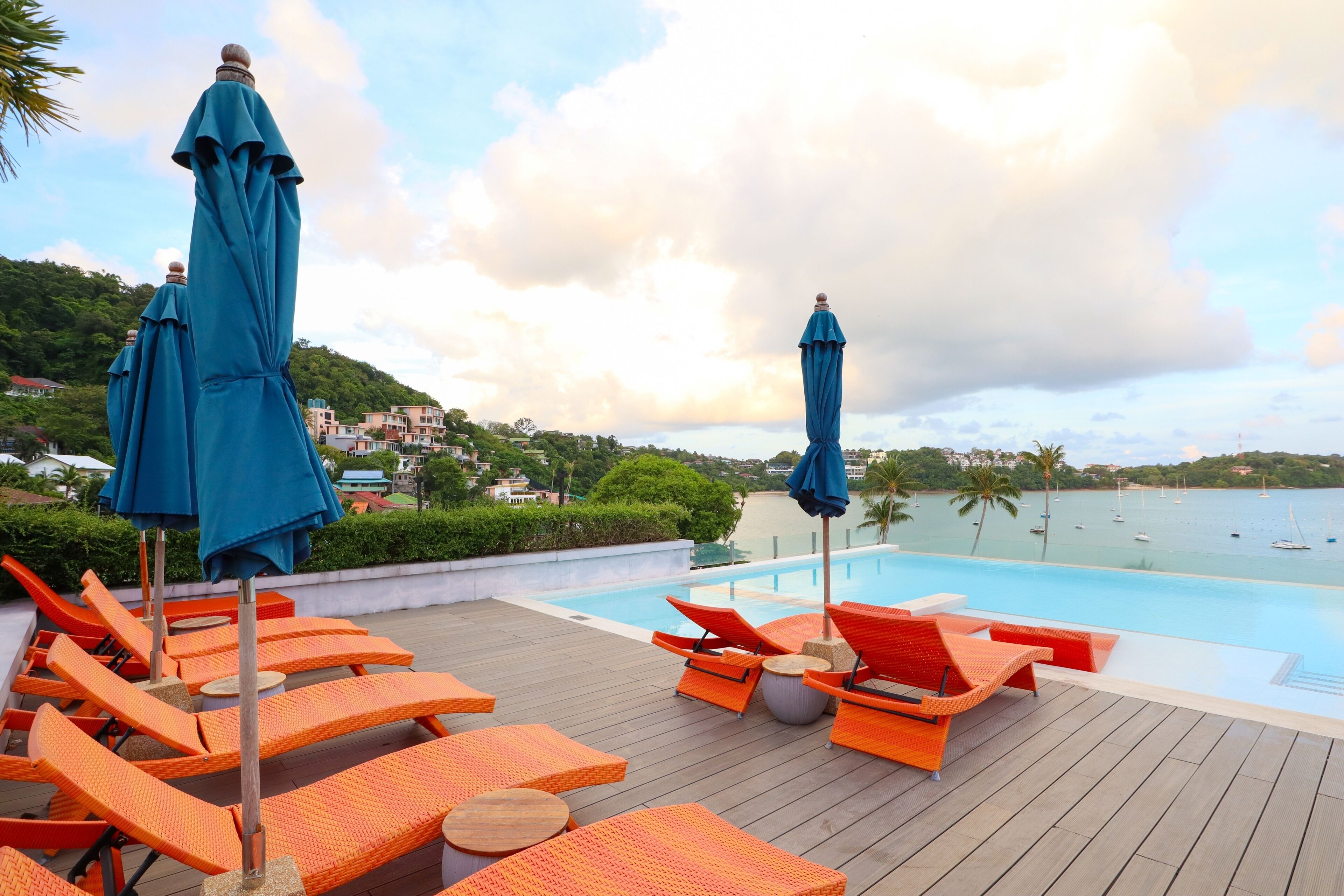 A poolside area with orange lounge chairs and blue umbrellas, overlooking a body of water with boats.