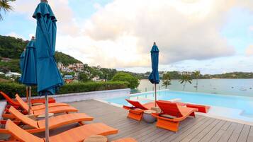 A poolside area with orange lounge chairs and blue umbrellas, overlooking a body of water with boats.