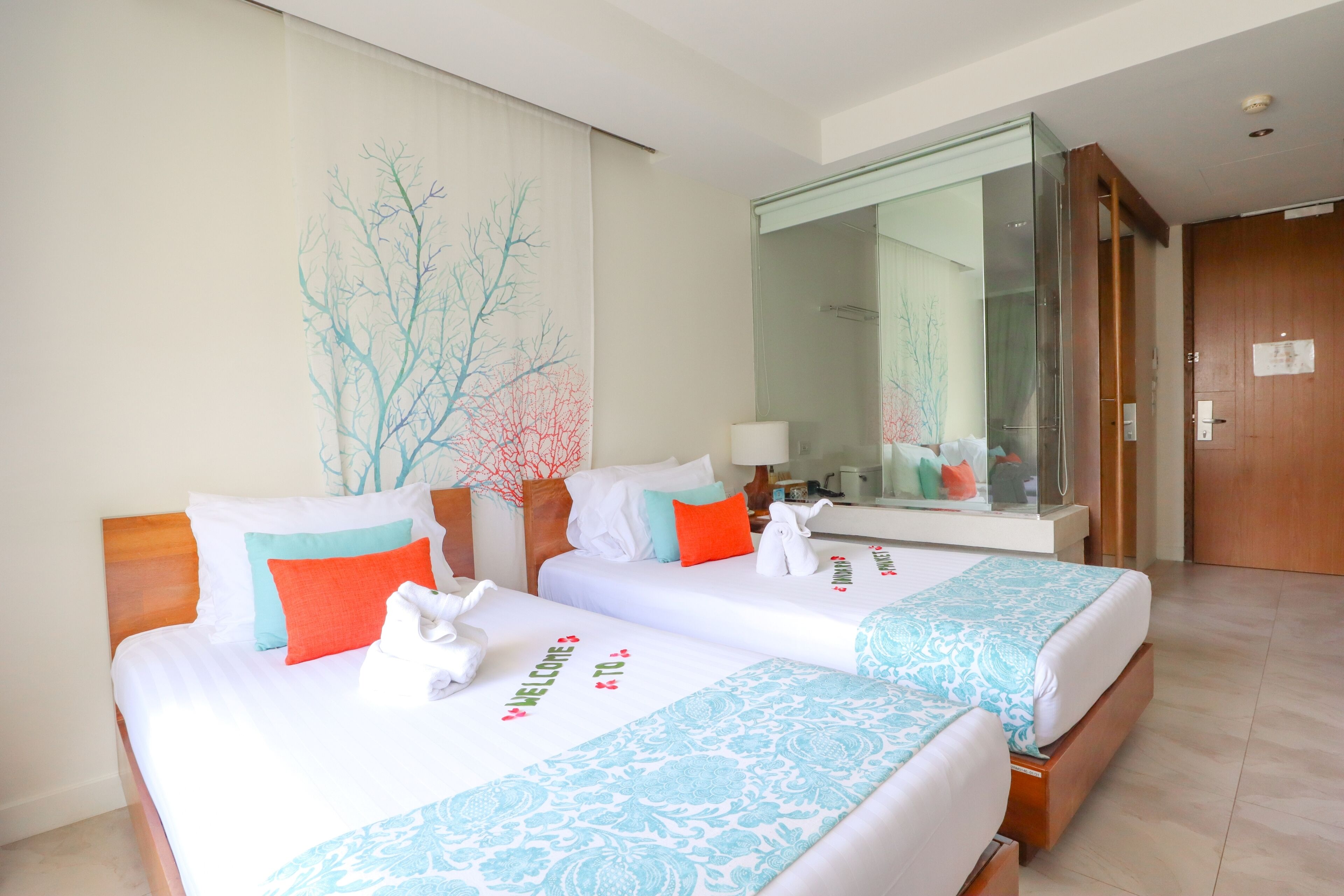A hotel room with two beds, each with a white bedspread and decorative pillows. There is a large mirror on one wall and a wooden door.