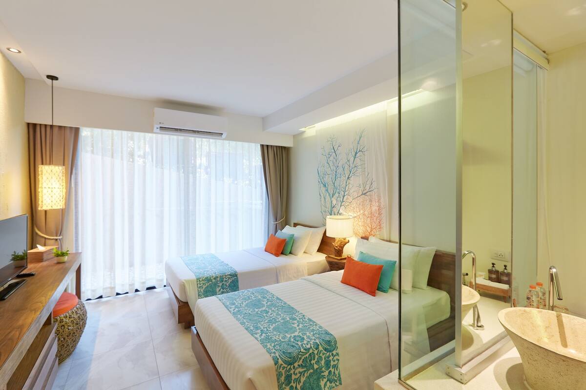 A hotel room with two beds, a TV, a desk, a large window with curtains, a wall-mounted air conditioner, and a bathroom with a sink and mirror.