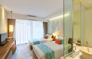 A hotel room with two beds, a TV, a desk, a large window with curtains, a wall-mounted air conditioner, and a bathroom with a sink and mirror. - Bandara Beach Resort, Phuket (Wichit)