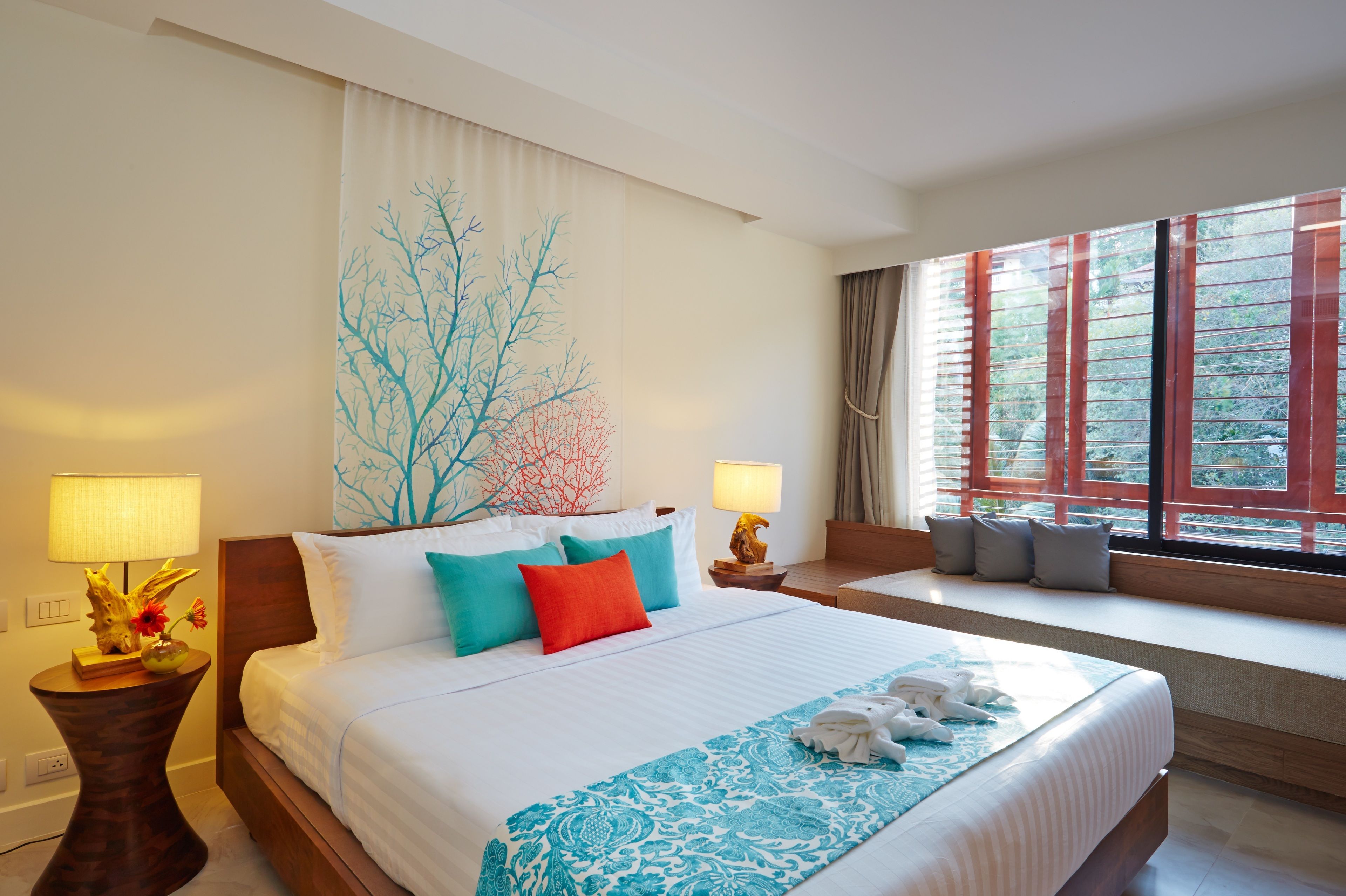 Ein Hotelzimmer mit einem großen Bett, einem Fenster mit Jalousien, einem Nachttisch mit Lampe und einer Wand mit einem Baum-Wandbild.