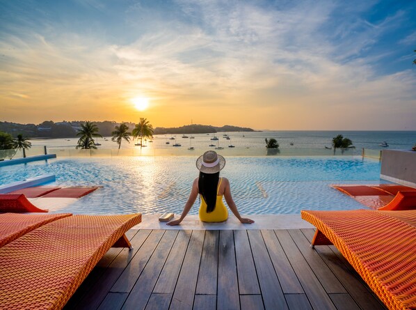 A woman sitting by an infinity pool with a sunset view, surrounded by lounge chairs. - Bandara Beach Resort, Phuket (Wichit)