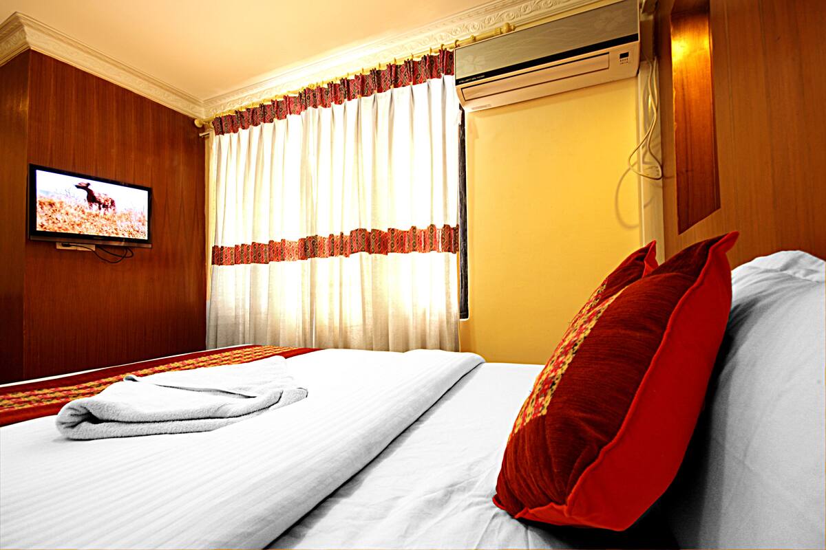 A hotel room with a bed, a television on the wall, and a window with curtains.