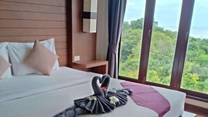 Ein Hotelzimmer mit einem großen Bett, Blick ins Grüne durch das Fenster und einem ordentlich bezogenen Bett mit Handtuch und Herz-Dekoration.