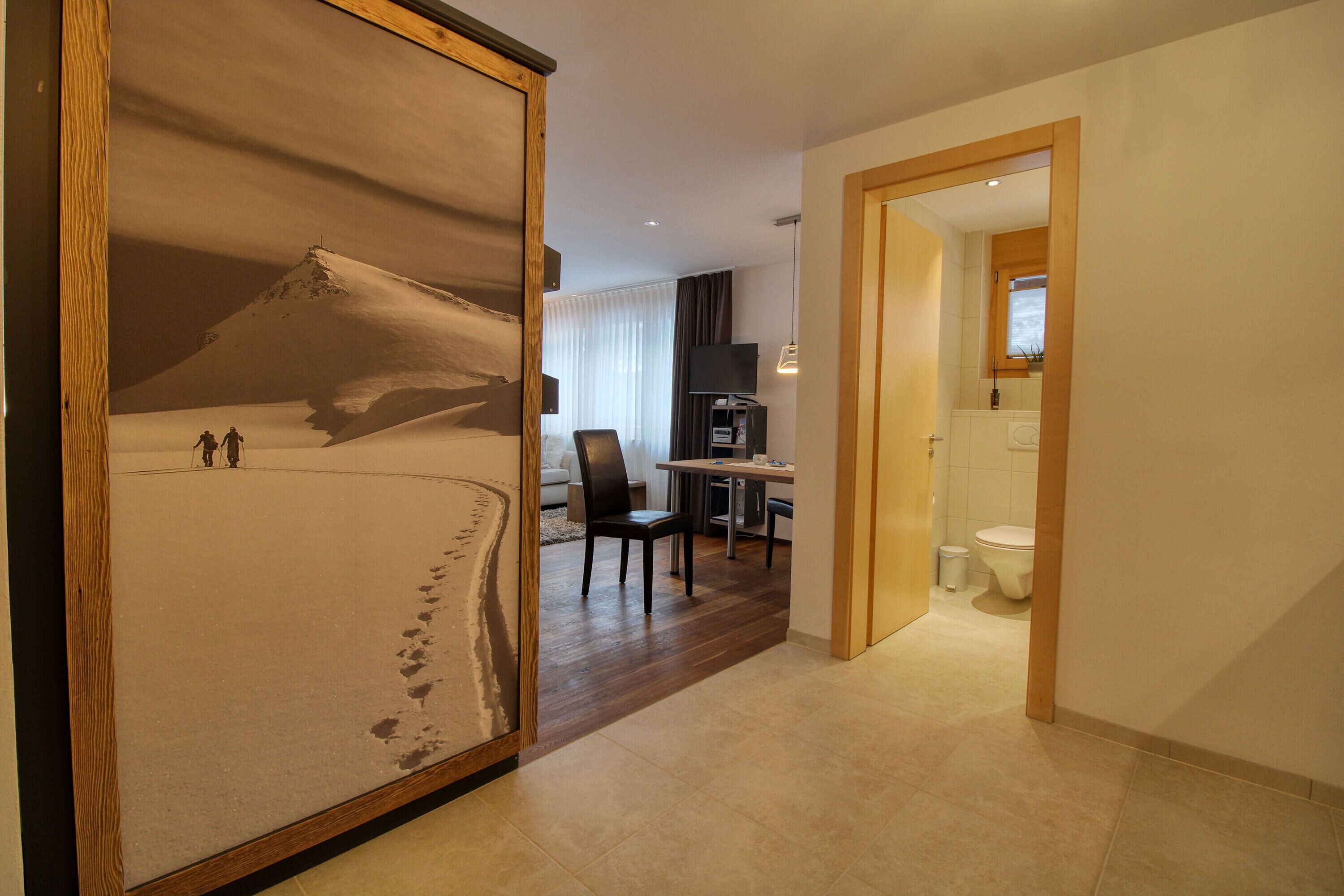 A hotel room with a large painting of a snowy mountain, a wooden floor, a dining area with a table and chairs, and a bathroom with a toilet and sink.