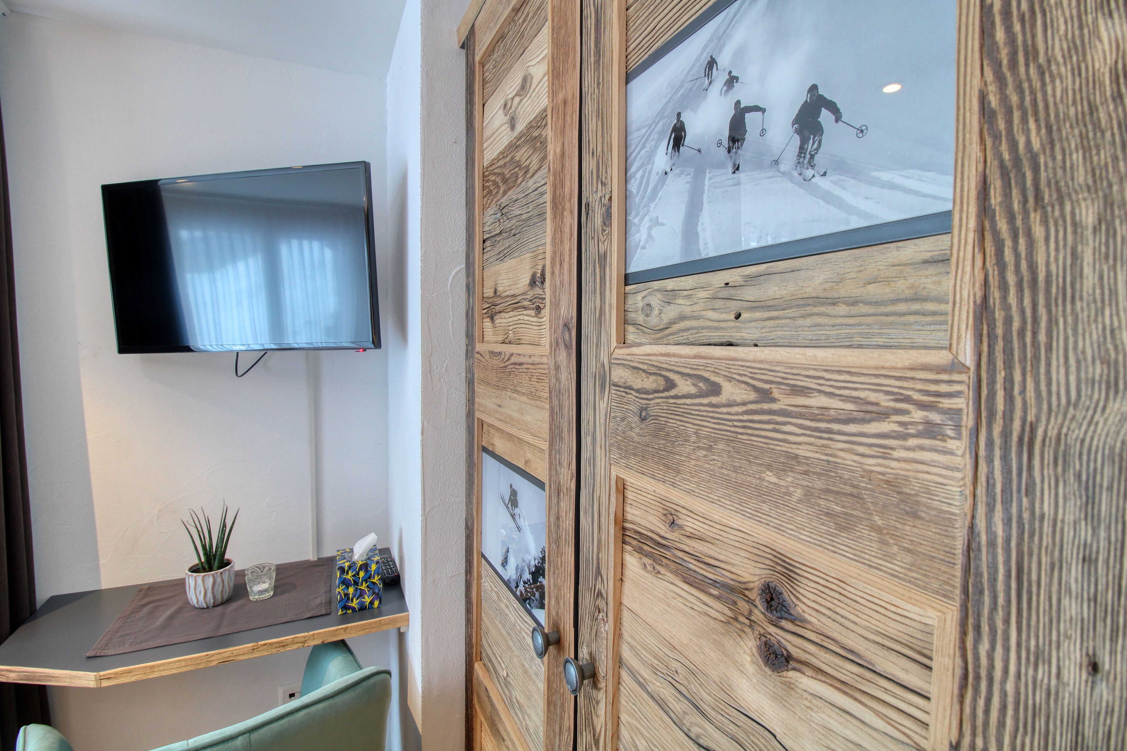 A room with a mounted TV, a wooden cabinet with a ski-themed glass door, a small plant, and a glass on a wooden table.