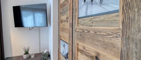 A room with a mounted TV, a wooden cabinet with a ski-themed glass door, a small plant, and a glass on a wooden table.