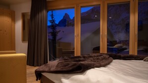 A bedroom with a bed, a wooden chair, a large window with a mountain view, and a sliding glass door. - Chalet A la Casa (Zermatt)