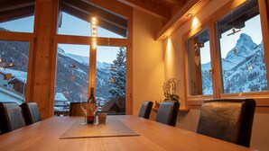 A wooden dining table with chairs, large windows with curtains, and a view of snowy mountains. - Chalet A la Casa (Zermatt)