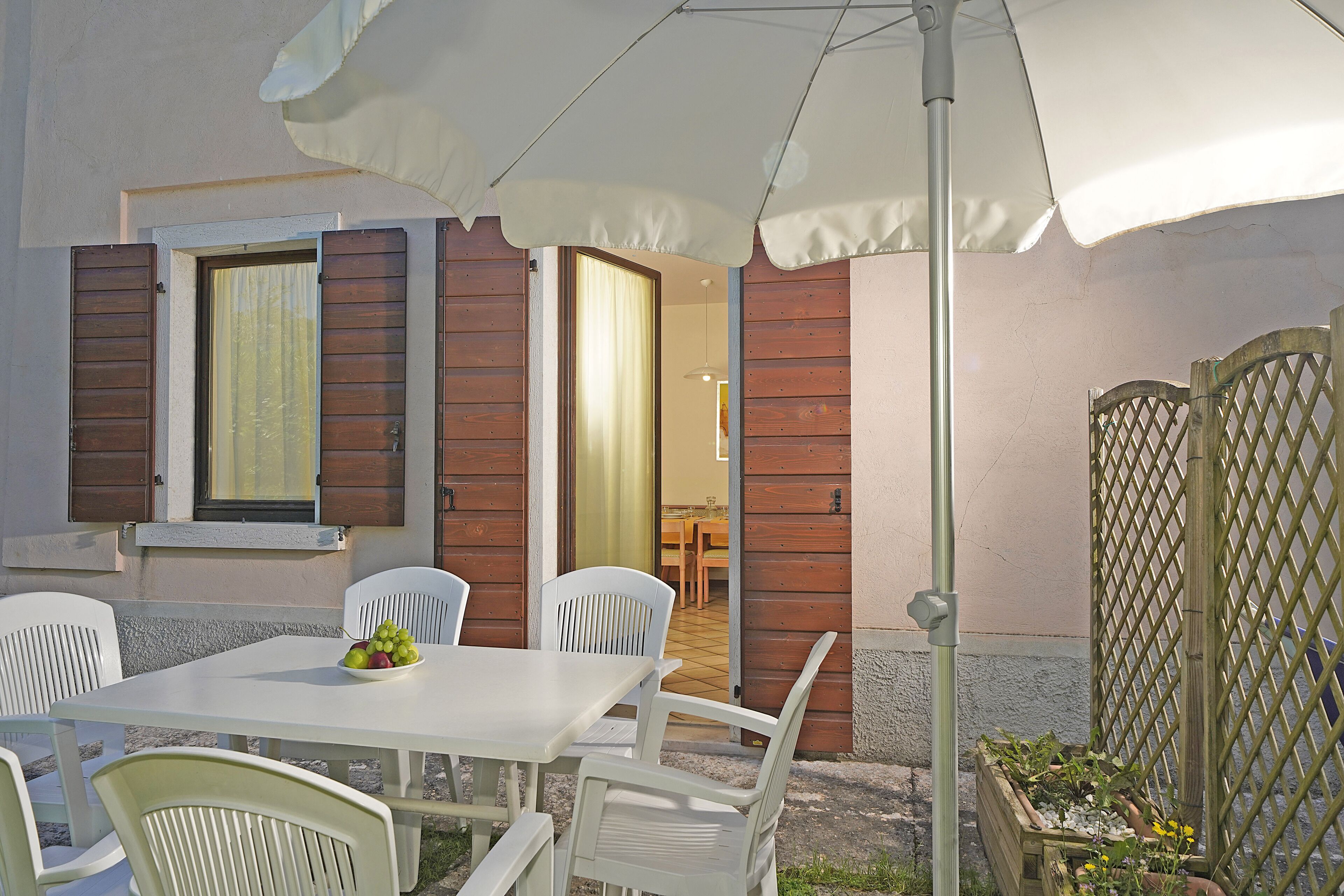 Eine Terrasse im Freien mit Tisch, Stühlen, einem weißen Sonnenschirm und Holzjalousien.
