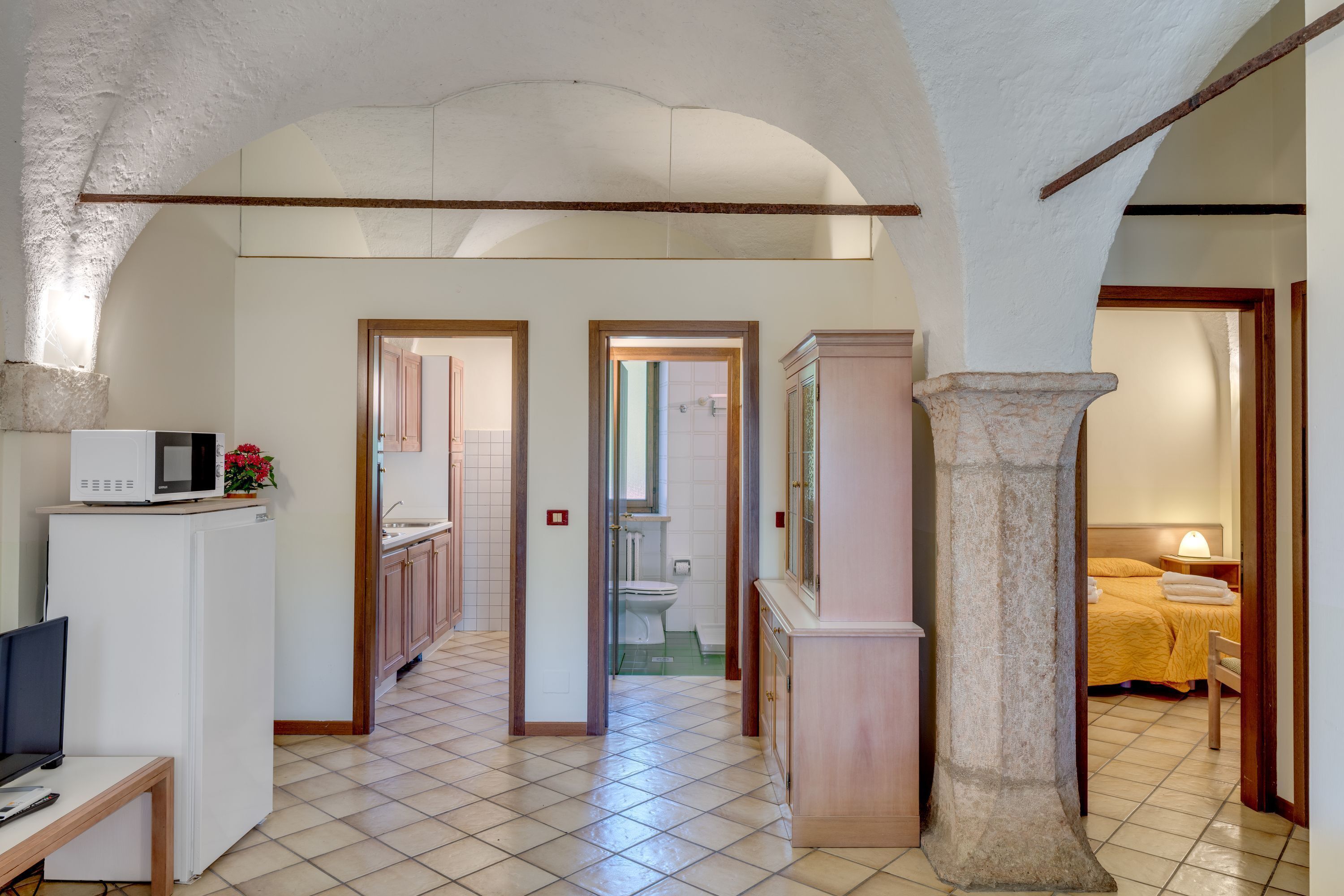 Un vaste couloir au sol carrelé mène à une salle de bain et à une chambre. Une grande colonne en pierre est visible au centre.