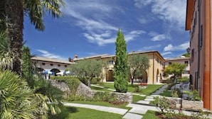 A courtyard with a stone pathway, palm trees, and buildings with arched windows. - Residence La Filanda (Costermano sul Garda)