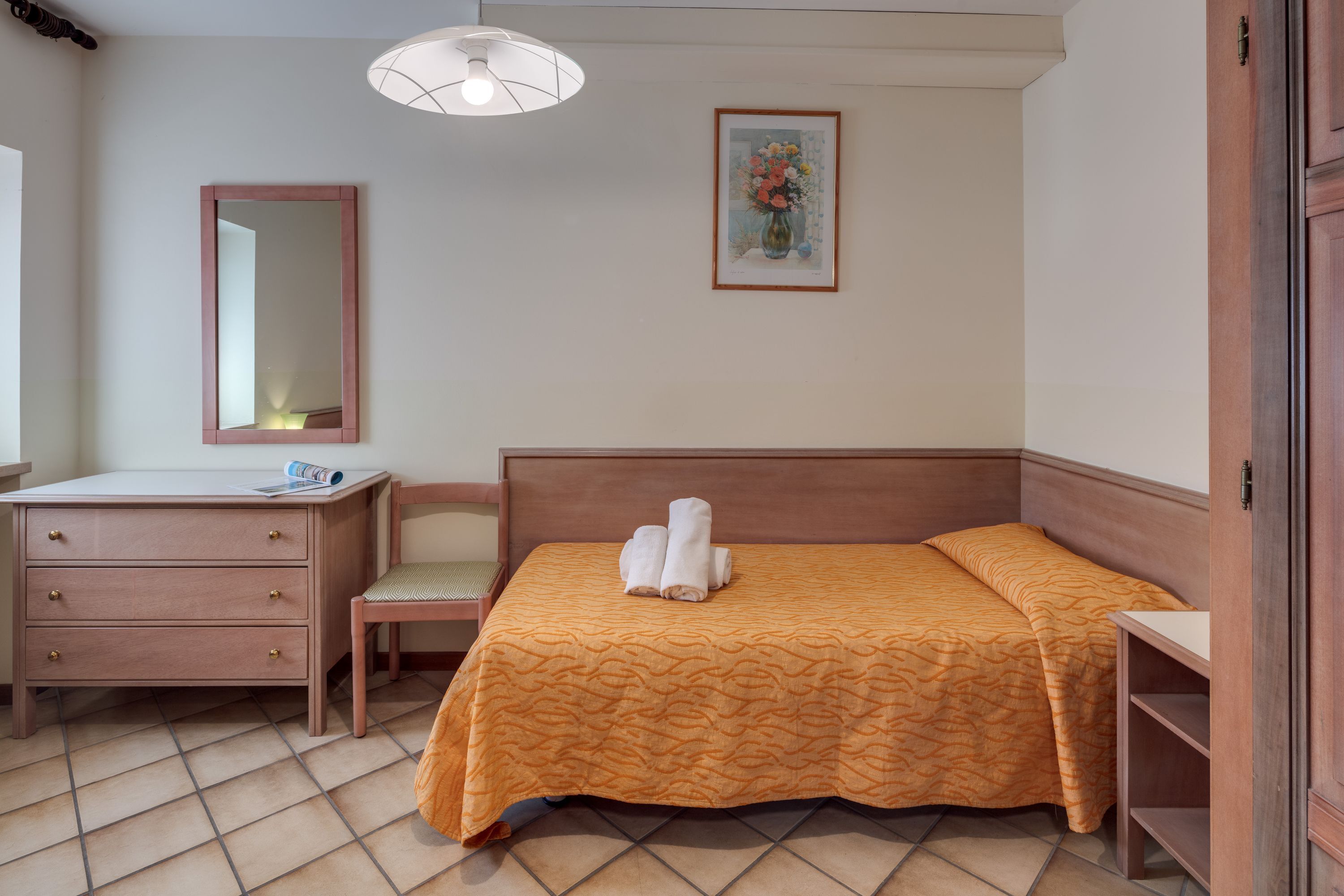 A hotel room with a bed, a wooden dresser, a chair, a desk, a mirror, and a painting on the wall.
