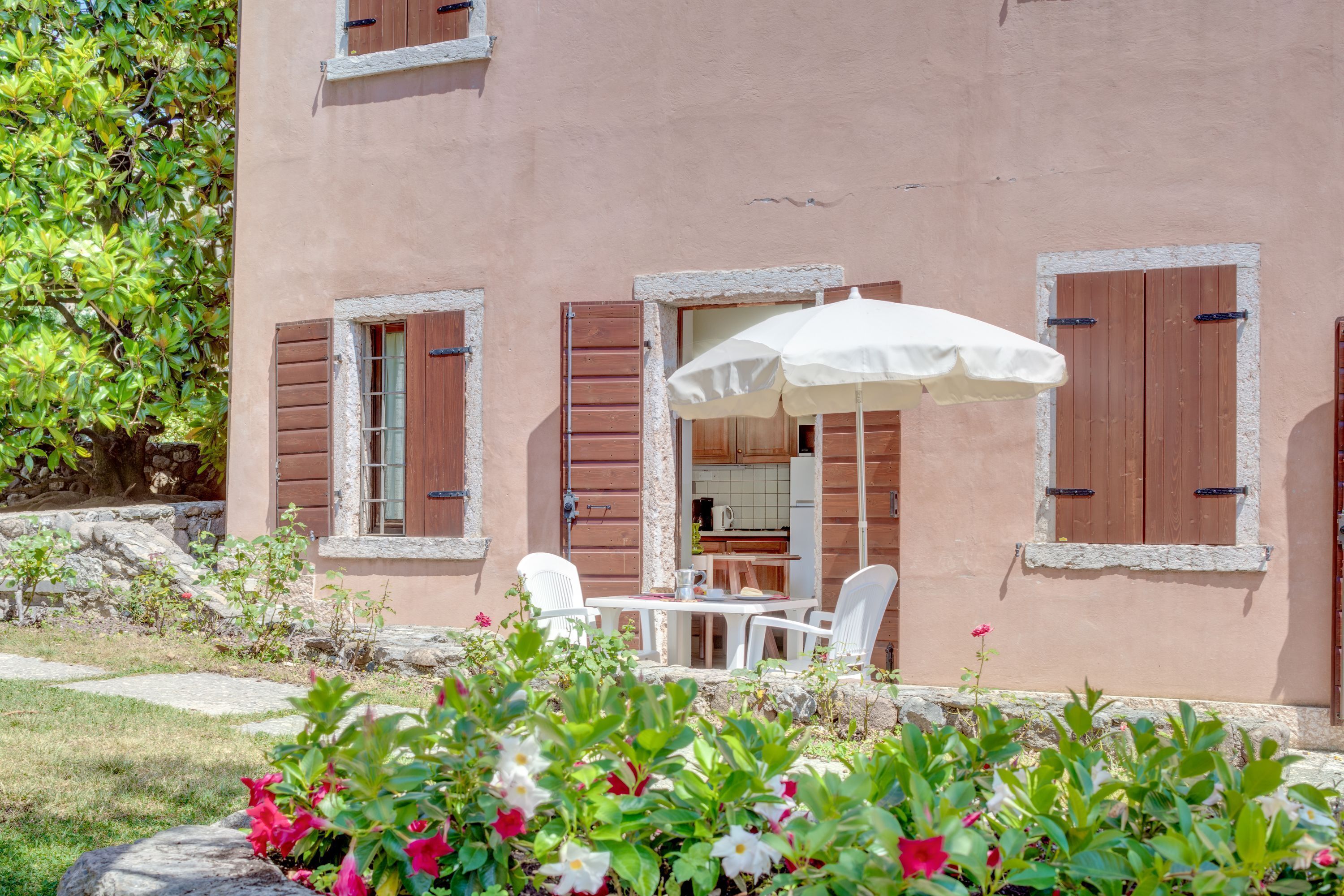 Ein Gebäude mit steinernen Fundament, einer durchgehenden Tür erkennbaren Küche und einer Terrasse mit Tisch und Stühlen unter einem weißen Sonnenschirm.