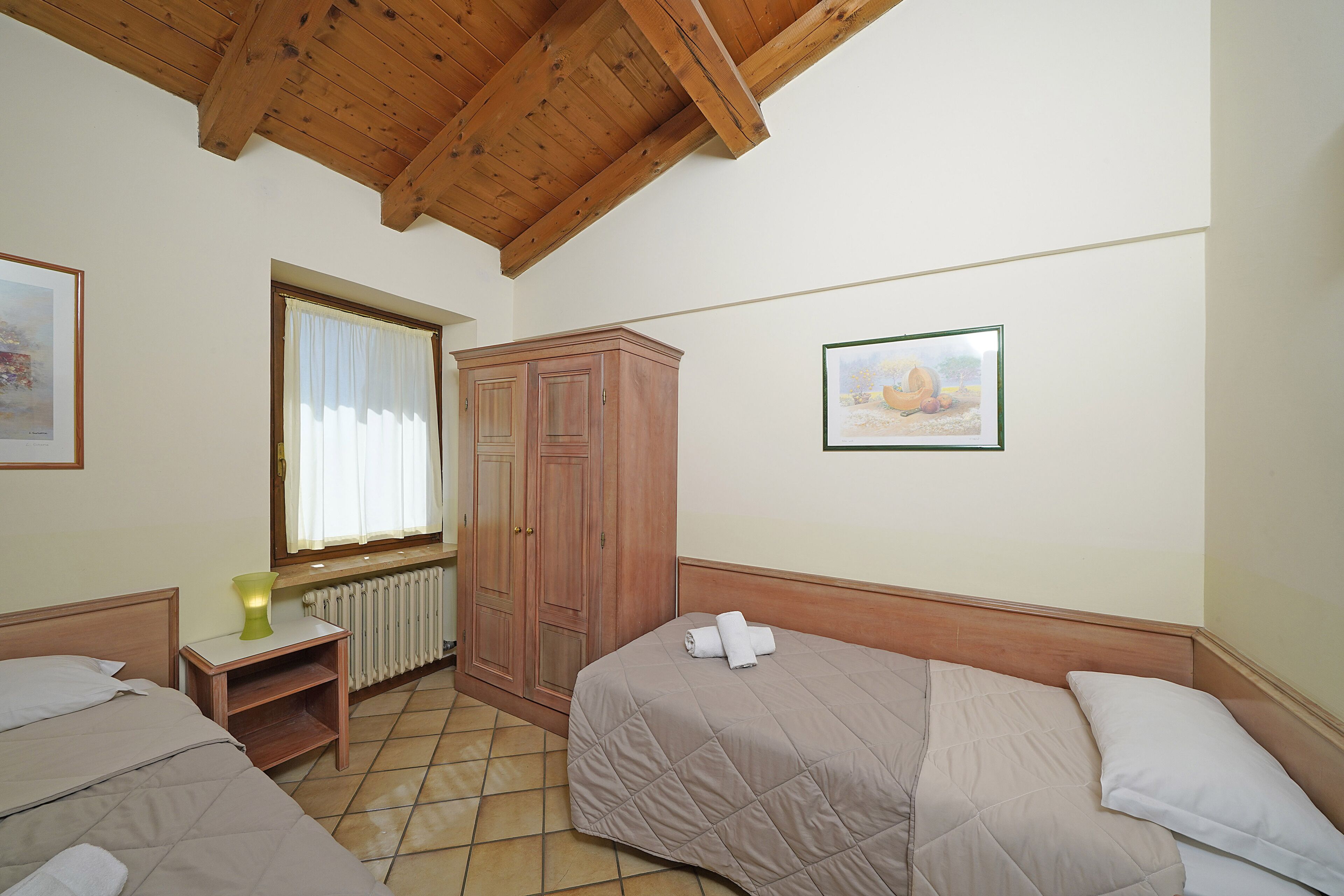 A bedroom with two beds, wooden headboards, a window with curtains, a radiator, a wooden wardrobe, and a framed picture on the wall.