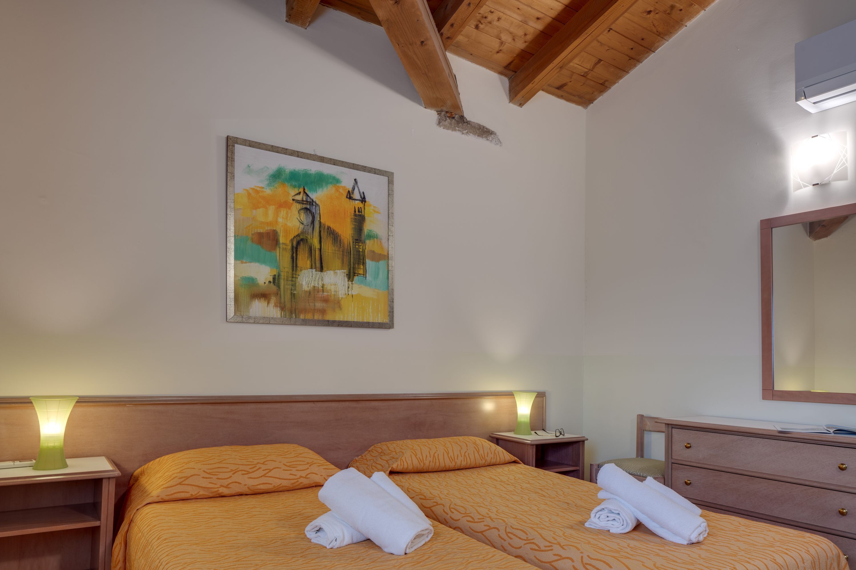 A hotel room with a wooden ceiling, a bed with orange bedding, a nightstand with a lamp, a dresser, and a painting of a cityscape on the wall.