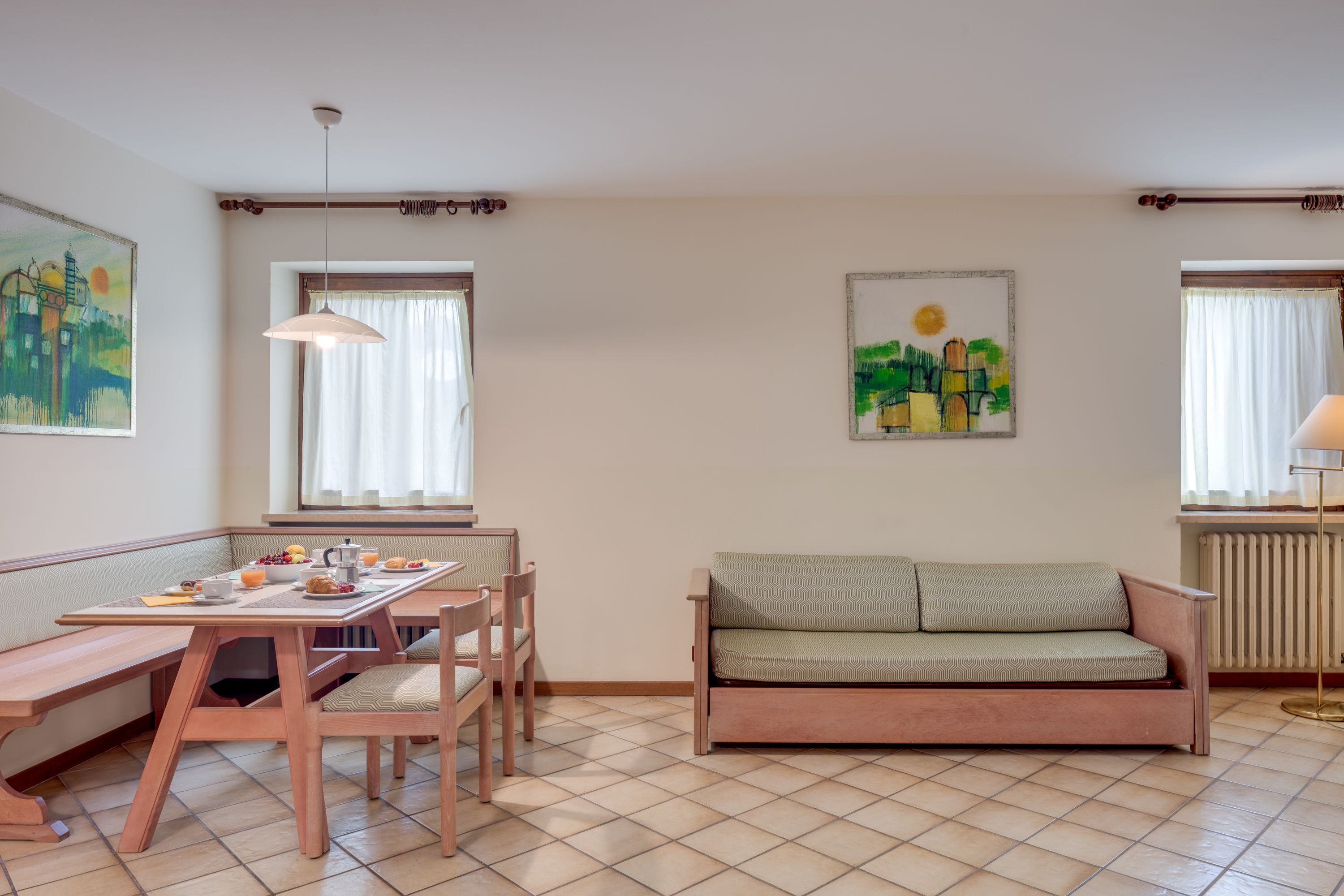 Un salon chaleureux avec un banc en bois, un canapé, une table à manger avec des chaises et deux fenêtres avec des rideaux.