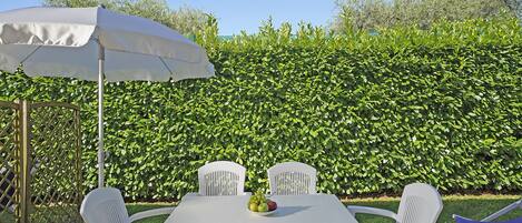 Una mesa blanca al aire libre con cuatro sillas y una sombrilla blanca, frente a un seto verde exuberante.