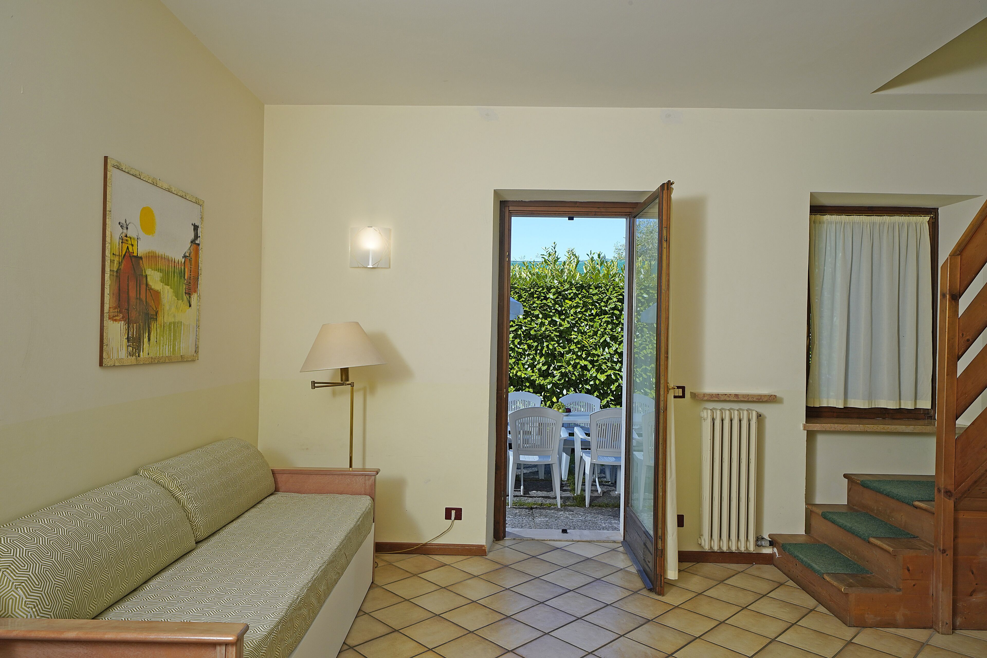 Une pièce comprenant un canapé, une lampe, un tableau, une porte donnant sur un espace extérieur avec une table et des chaises, ainsi qu’un escalier.