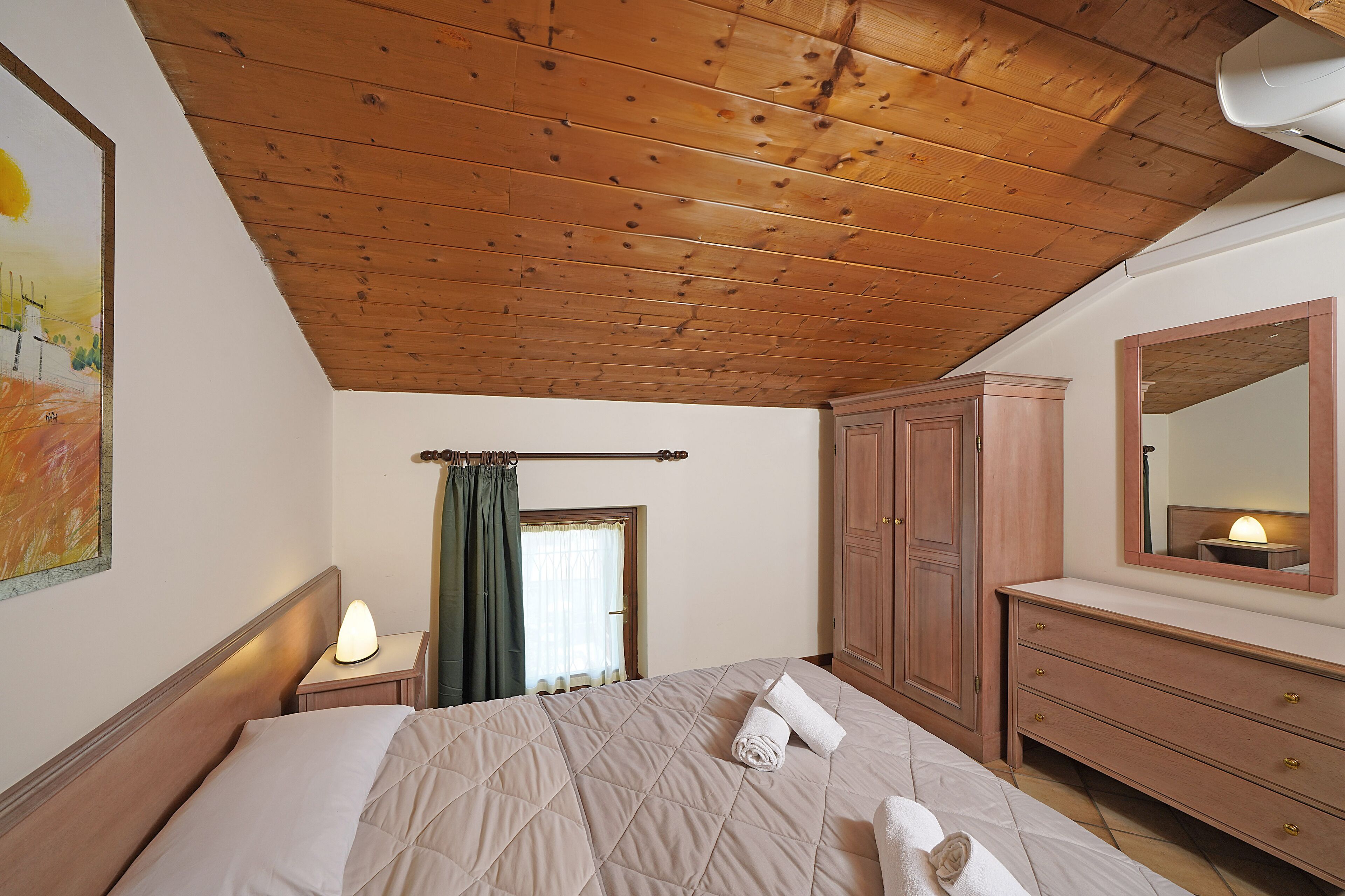 Um quarto com teto de madeira, uma cama com colcha, uma mesa de cabeceira com abajur, cômoda de madeira e espelho na parede.