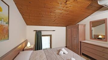 Um quarto com teto de madeira, uma cama com colcha, uma mesa de cabeceira com abajur, cômoda de madeira e espelho na parede.