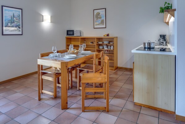 A dining area with a wooden table and chairs, a kitchen counter with appliances, and shelves with dishes and a microwave. - Appartamenti San Carlo (Costermano sul Garda)