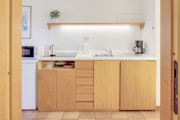 A compact kitchen with a microwave, kettle, coffee maker, and a shelf above the counter.