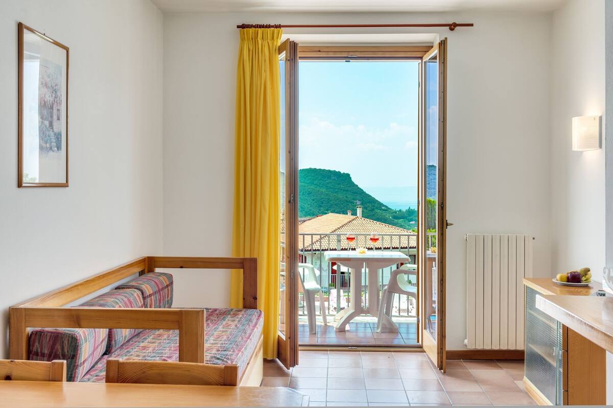 A room with a bed, a dining table, and a view of a balcony with outdoor furniture.
