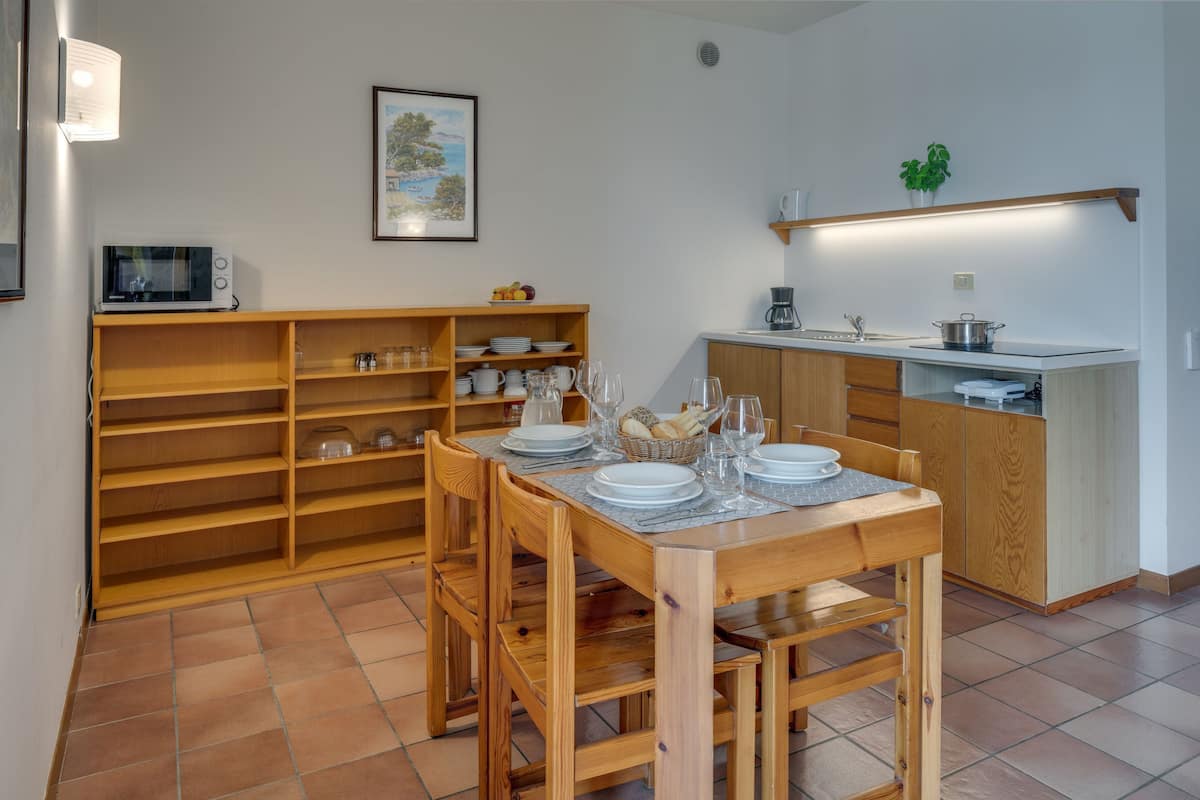 A dining area with a wooden table set for four, surrounded by wooden chairs. A kitchen with a white countertop and wooden cabinets is visible in the background.