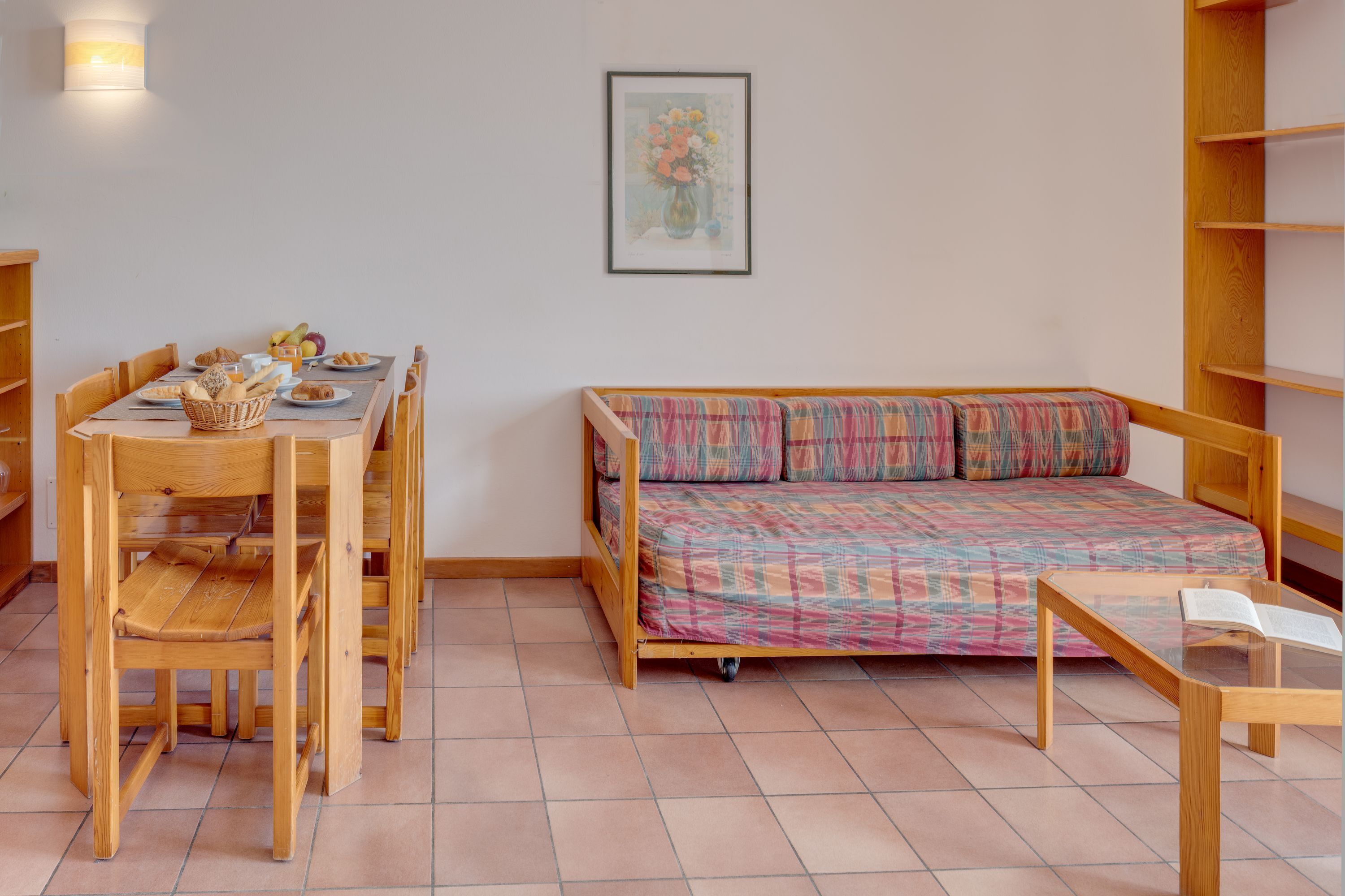 A room with a wooden bed, a dining table with chairs, and a small table with an open book.